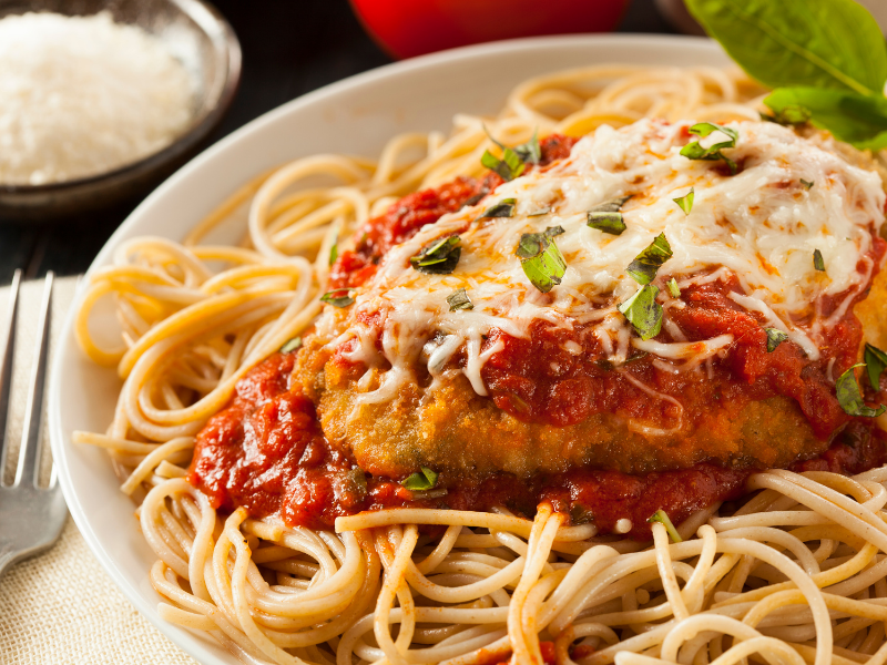 Chicken parmesan in a white plate coated in breadcrumbs with tomato sauce and parmesan cheese melted on top over spaghetti noodles with parmesan cheese on the side